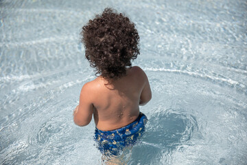 Cute boy two year old toddler with brown curly hair playing in a resort or hotel swimming pool on a beautiful sunny day as seen from back angle isolated against water.