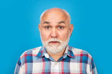 Portrait of an surprised bald old mature senior man with grey beard in shirt isolated on blue studio background.