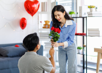 Millennial Asian young romantic lover couple male boyfriend kneel down giving red roses bouquet surprising female girlfriend celebrating anniversary on valentine day festival in decorated living room