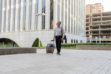 Senior businessman with suitcase, male professionals in suit walking on sidewalk next to business office building starting business trip. - Powered by Adobe