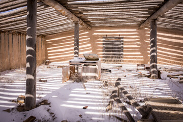 Fototapeta na wymiar Interior of abandoned adobe potato bunker/storage barn in winter. Heavy beams, snow on ground, sunlight casting strong shadows on entire room. Abandoned appliance and other artifacts.