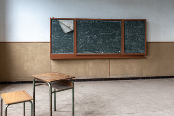 Abandoned and run-down public accessible classroom with scribbles left on the blackboard and wooden table with chair