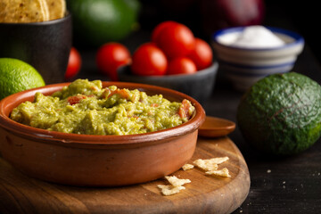 Close-up of bowl with guacamole on dark table with tomatoes, avocados, limes and salt, selective focus, horizontal