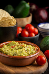 Close-up of bowl with guacamole on dark table with nachos, tomatoes, avocados and salt, selective focus, black background, vertical