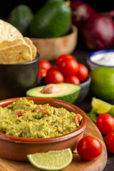 Top view of bowl with guacamole on dark table with nachos, tomatoes, avocados, sliced lime and salt, selective focus, vertical