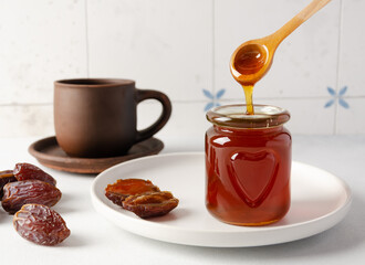 Natural organic healthy date syrup flows from wood spoon into a glass jar with whole dates and a...
