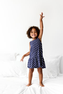 Happy little girl with afro hair standing on white bed raising hand