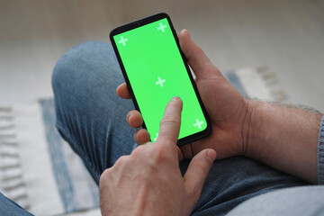 Young man sitting at home holding smartphone green mock-up screen in hand. Male person using chroma...
