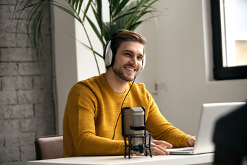 Man blogger smiling while broadcasting his live audio podcast in studio using headphones, laptop...