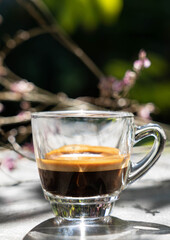 Espresso shot coffee in clear small cup with blur pink flower in background in the morning