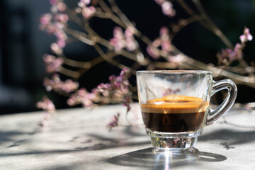 Espresso shot coffee in clear small cup with blur pink flower in background in the morning