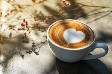 Obraz na płótnie Canvas Cup of heart shape latte art coffee with shadow of tree branch and some dry flower behind on desk in the morning