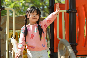 Happy asian girl having fun on playground in public park surrounded by green trees at sunlight morning