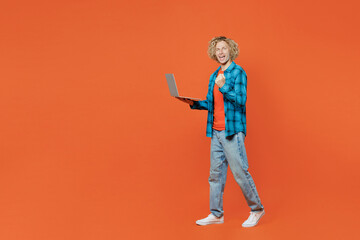 Full body young blond caucasian IT man wear blue shirt orange t-shirt hold use work on laptop pc computer do winner gesture isolated on plain red background studio portrait. People lifestyle concept.