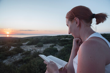 kobieta czytająca książkę nad brzegiem morza o zachodzie słońca