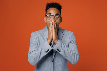 Young surprised amazed successful employee business man corporate lawyer in classic formal grey suit shirt glasses cover mouth with hand work in office isolated on plain red orange background studio