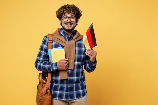 Young fun happy teen Indian boy IT student he wears casual clothes shirt glasses bag hold in hands books German flag isolated on plain yellow color background High school university college concept.
