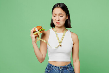 Young addicted sad woman with tied hand neck by measure tape wears white clothes hold eat burger isolated on plain pastel green background. Proper nutrition healthy fast food unhealthy choice concept.