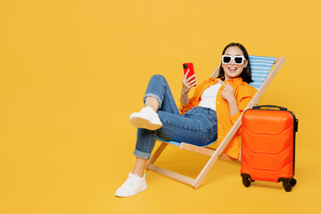 Young woman wear summer clothes sit in deckchair use show mobile cell phone isolated on plain yellow background. Tourist travel abroad in free spare time rest getaway. Air flight trip journey concept.