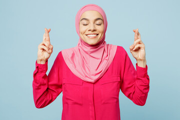 Young arabian asian muslim woman in pink abaya hijab keep finger crossed make wish, eyes closed isolated on plain pastel light blue cyan background studio portrait People uae islam religious concept