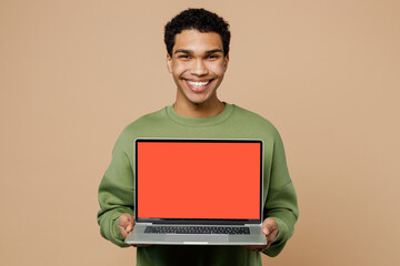 Young IT man of African American ethnicity wear green sweatshirt hold use work on laptop pc computer with blank screen workspace area isolated on plain pastel light beige background studio portrait.