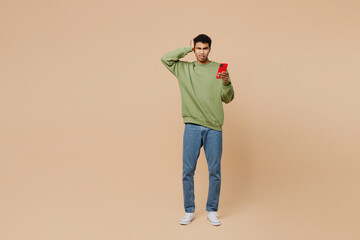 Fototapeta premium Full body sad young man of African American ethnicity wear green sweatshirt hold scratch head use mobile cell phone isolated on plain pastel light beige background studio . People lifestyle concept.