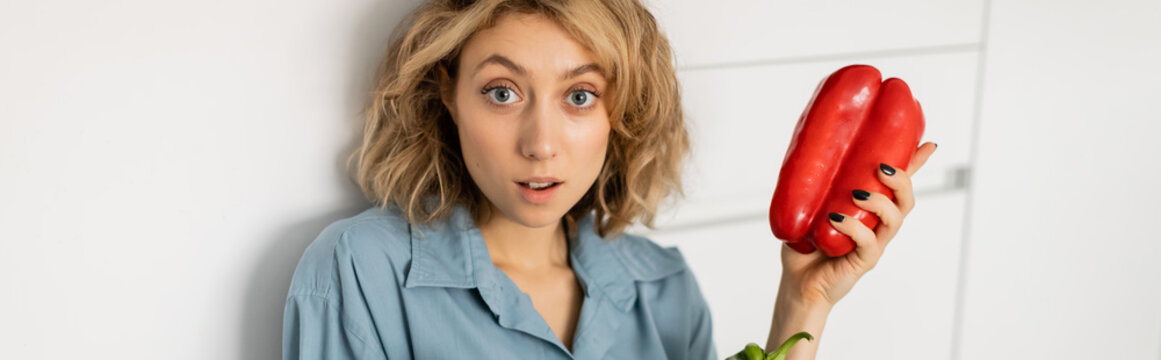 shocked young woman with wavy hair holding red bell pepper in kitchen, banner.