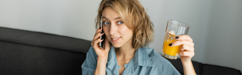 cheerful woman with wavy hair holding glass of orange juice while talking on smartphone, banner.