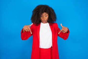 Amazed young businesswoman with afro hairstyle wearing red over blue background points down with fore fingers, opens mouth being shocked. Advertisement concept.