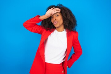 young businesswoman with afro hairstyle wearing red over blue wall wiping forehead with hand making...