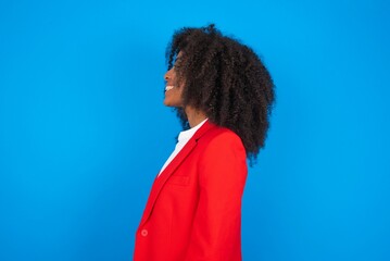 Profile of smiling young businesswoman with afro hairstyle wearing red over blue background with healthy skin, has contemplative expression, ready to have outdoor walk.