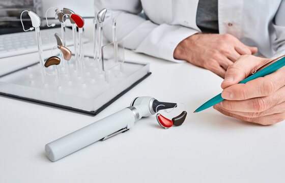 Audioprosthetist presentation hearing aids in two colors to his patient lying on table near otoscope and display stand with BTE hearing aids