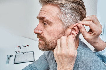 Audiologist inserting ITE hearing aid in adult man's ear at audiology center, close-up, side view. Deafness treatment, hearing solutions