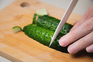Female fingers with a knife cut a cucumber, close-up, on a wooden cutting board
