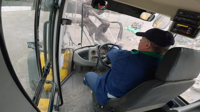 Inside cab view of a man driving heavy machine vehicle, a front loader grabbing garbage and loading a truck, fast motion. Waste management concept.