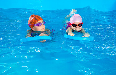 Two little girls learning how to swim in swimming pool using flutter boards