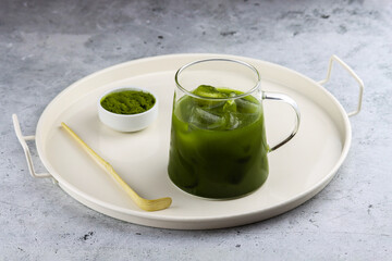 Matcha green tea with ice cubes on a serving tray with a small bowl on matcha powder and matcha spoon. Grey background. Iced matcha. Antioxidant organic drink made of ground powder of green tea leaves