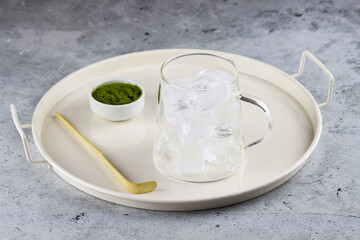 Serving plate with a set for preparing matcha green tea. A small bowl of matcha powder, matcha spoon and a transparent glass filled with ice cubes for matcha latte antioxidant invigorating drink