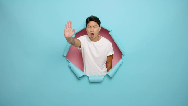 Young strict man 20s in casual white t-shirt looking camera showing stop gesture with palm refusing, isolated next to blue torn color background studio portrait. People lifestyle concept