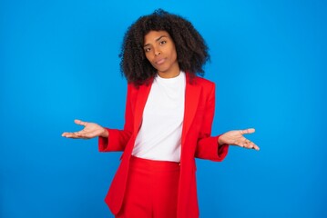 young businesswoman with afro hairstyle wearing red over blue background looks uncertain shrugs...