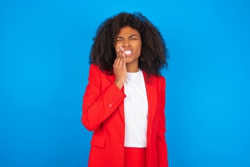 Tooth ache concept. young businesswoman with afro hairstyle wearing red over blue, feeling pain,...