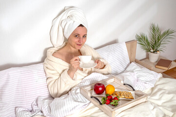 Woman eating fruit breakfast in bed at home in the morning. Lady wrapping a towel around head. Girl wearing bathrobe on clean white bedding with cozy blanket. Healthy lifestyle