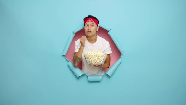 Asian man 20s wearing white t-shirt holding big plate with popcorn while looking at camera, looks a movie with different spectrum of emotions, isolated next to blue torn background