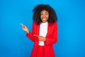 young businesswoman with afro hairstyle wearing red over blue background points at copy space...