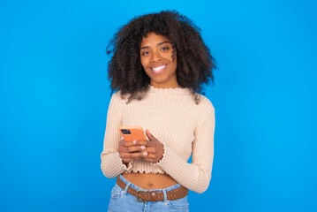 Young woman with afro hair style wearing crop top over blue background Mock up copy space. Using mobile phone, typing sms message