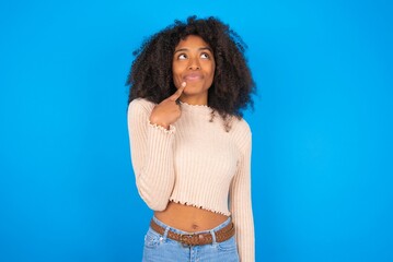 Lovely dreamy Young woman with afro hair style wearing crop top over blue background keeps finger...