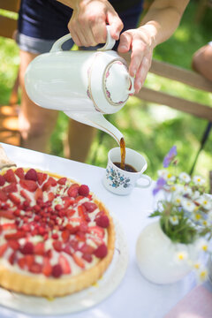 Kaffeetrinken im sommerlichen Garten mit Erdbeertorte. Eine Person in kurzer Hose gießt Kaffe aus einer schönen Porzellankanne in eine Blümchentasse.