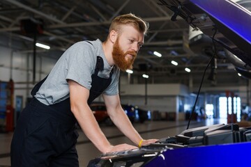 Fototapeta na wymiar A brutal bearded man in uniform works at a car repair station. Checking the car in a clean modern space