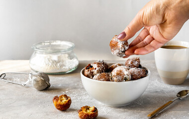 Italian traditional Carnival fritters toped with sugar powder in white bowl, woman hand is holding one of the sweets, light concrete background with cacao mug and sugar in jar