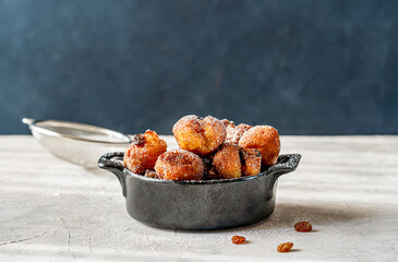 Carnival fritters or balls are Italian sweets dropped with sugar powder in a black pot on concrete table with dark background, strainer, strainer, raisins